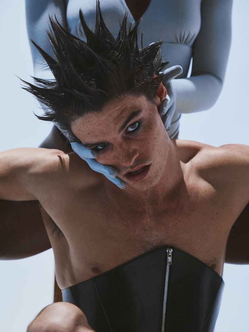 Striking studio portrait of a young adult male with a spiky hairstyle, wearing a black corset, with gloved hands touching his face.