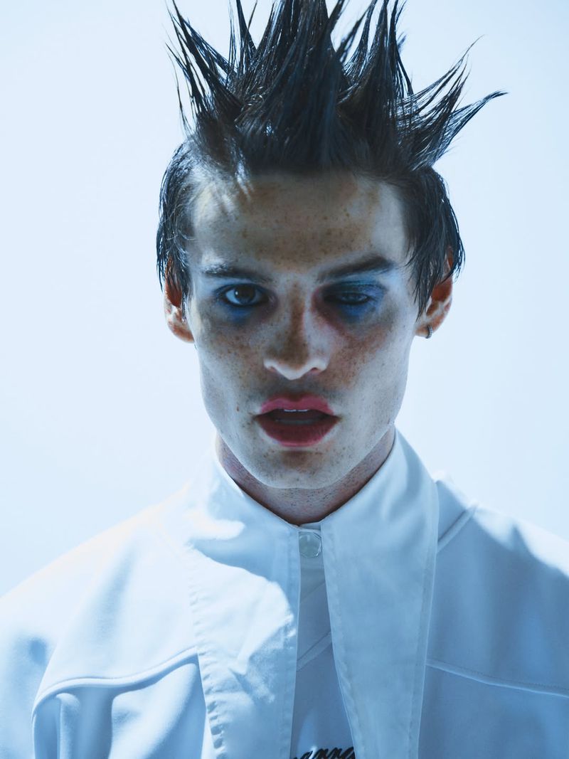 Close-up portrait of a male model with spiky dark hair, blue eye makeup, and white shirt against a bright background