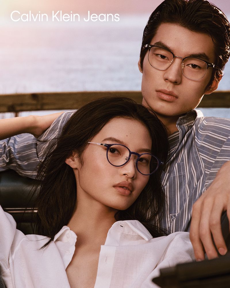 Two young adults pose in an outdoor setting. The woman, wearing glasses and a white top, is in the foreground. The man, wearing glasses and a striped shirt, is in the background.