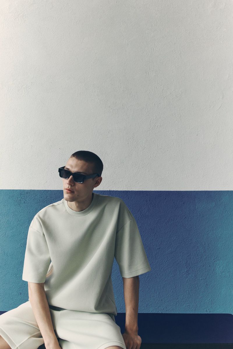 Model wearing dark sunglasses and beige t-shirt seated against a two-tone white and blue textured wall for H&M.