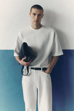 Male model in white t-shirt and white jeans holding a black bag against a two-tone wall for H&M man