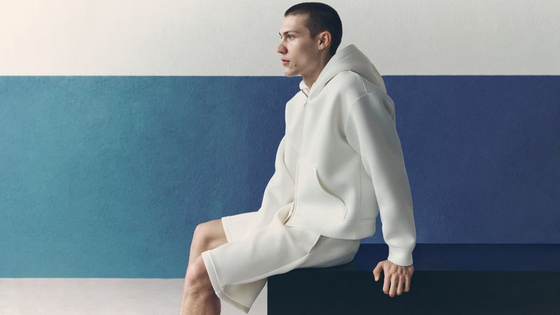 Model seated wearing an off-white hoodie and shorts against a two-tone blue and white wall for H&M man Summer Scuba.