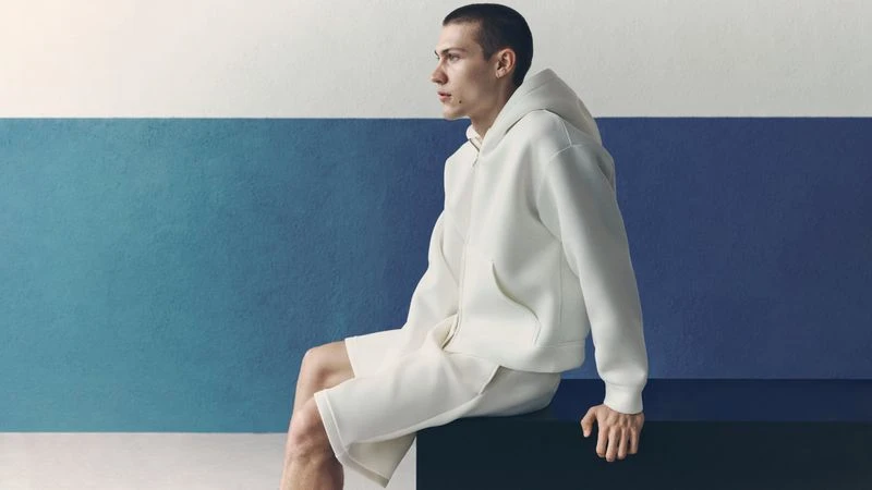 Model seated wearing an off-white hoodie and shorts against a two-tone blue and white wall for H&M man Summer Scuba.