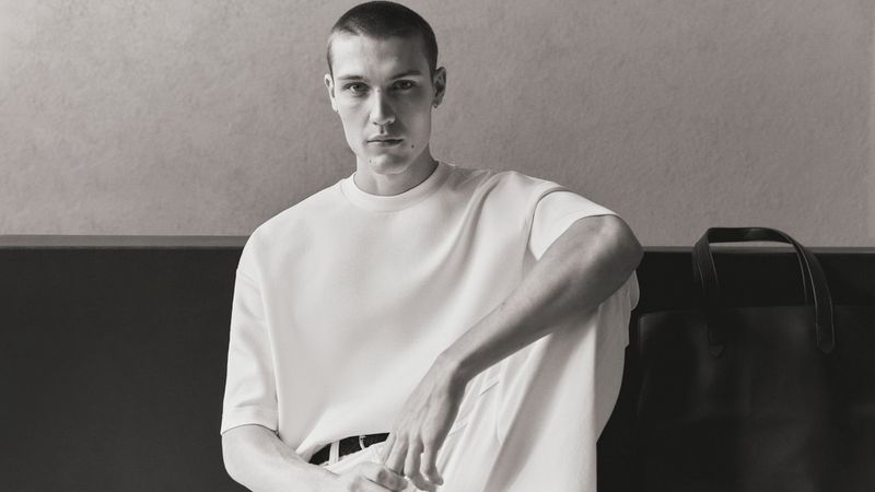 Monochrome portrait of a male model in a white t-shirt sitting beside a dark tote bag against a plain wall.