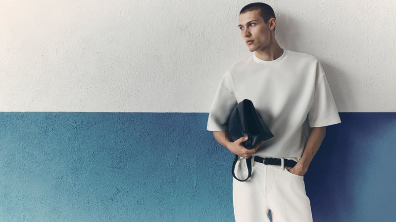 Model wearing a white t-shirt and white jeans holding a black bag against a split white and blue wall.