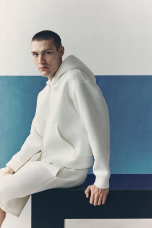Model wearing a white zip-up hoodie sitting against a white and blue wall for the H&M man Summer Scuba project.