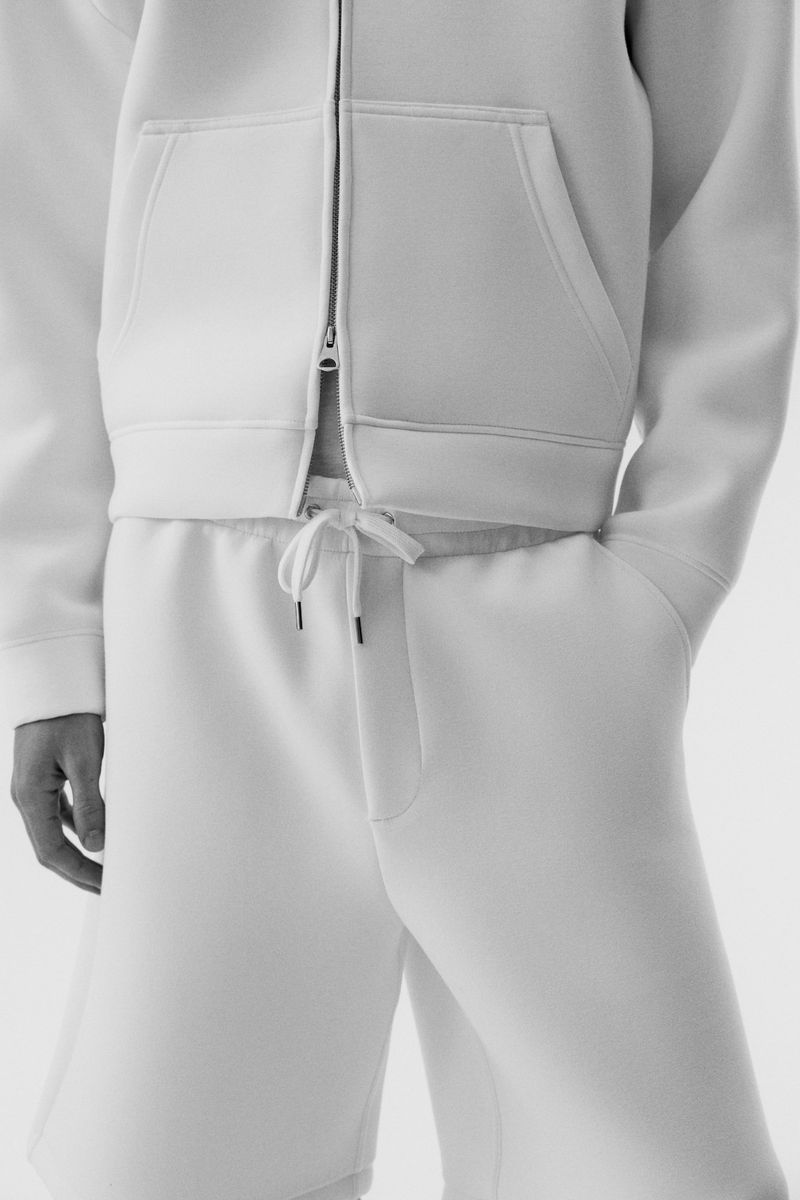 Close-up of a person wearing a white zip-up scuba hoodie and matching white shorts against a seamless white background.