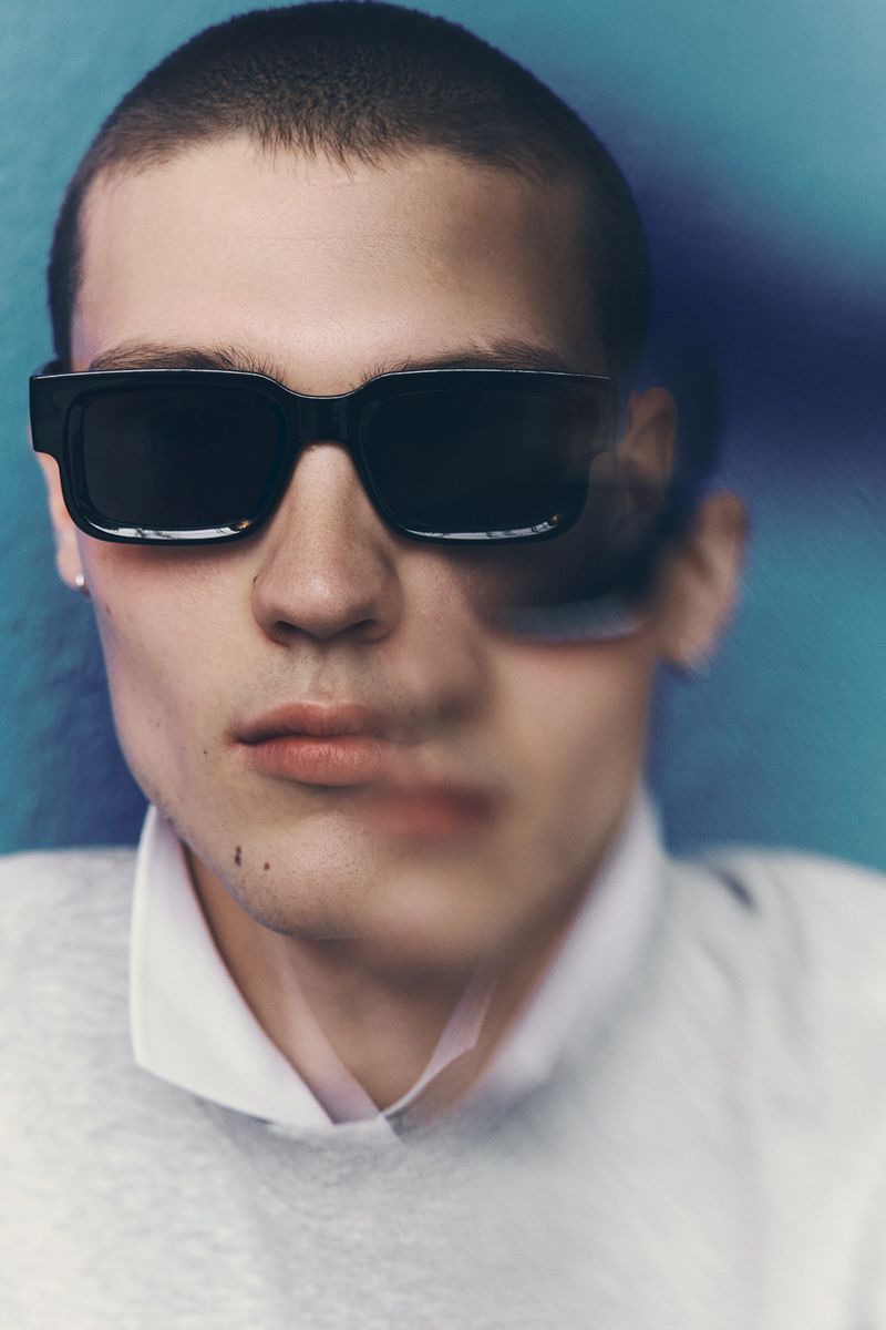 Close-up portrait of a young man wearing thick black sunglasses and a white collared shirt against a blue backdrop.