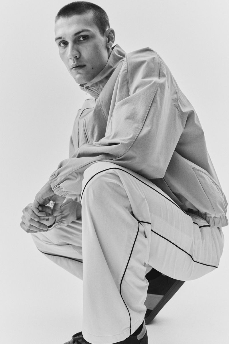 Monochrome portrait of a male model crouching, wearing a lightweight windbreaker jacket and matching trousers against a plain white background.