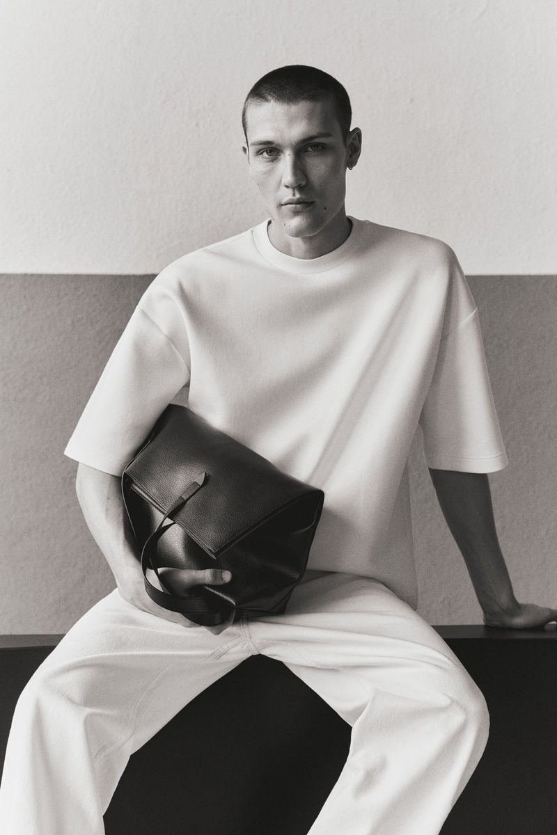Man wearing white t-shirt and white trousers holding a large black leather bag against a studio wall