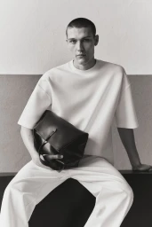 Man wearing white t-shirt and white trousers holding a large black leather bag against a studio wall