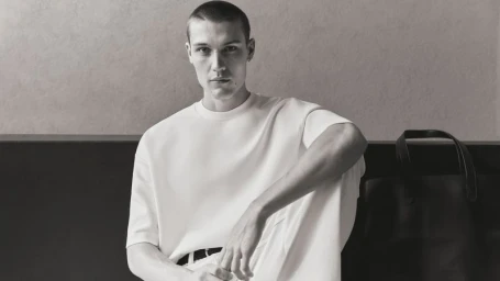 Monochrome portrait of a male model in a white t-shirt sitting beside a dark tote bag against a plain wall.