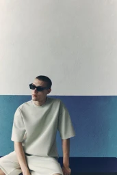 Model wearing dark sunglasses and beige t-shirt seated against a two-tone white and blue textured wall for H&M.