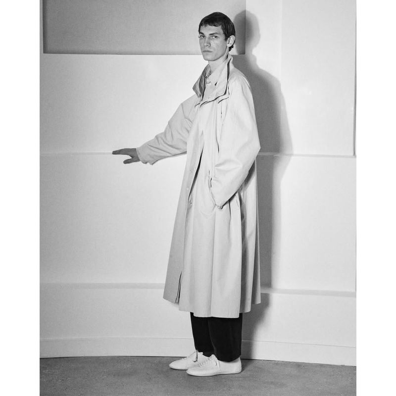 Monochrome portrait of a male model wearing an oversized coat standing against a white studio wall.