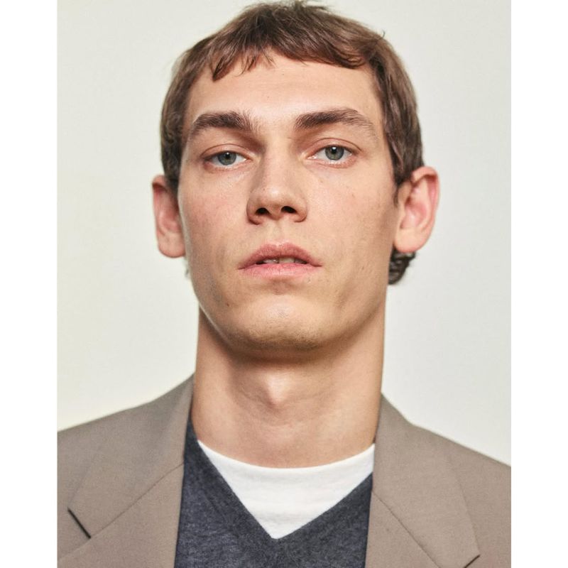Close-up of an adult male in a taupe blazer over a white t-shirt and grey v-neck sweater, posing in an indoor setting against a neutral background.