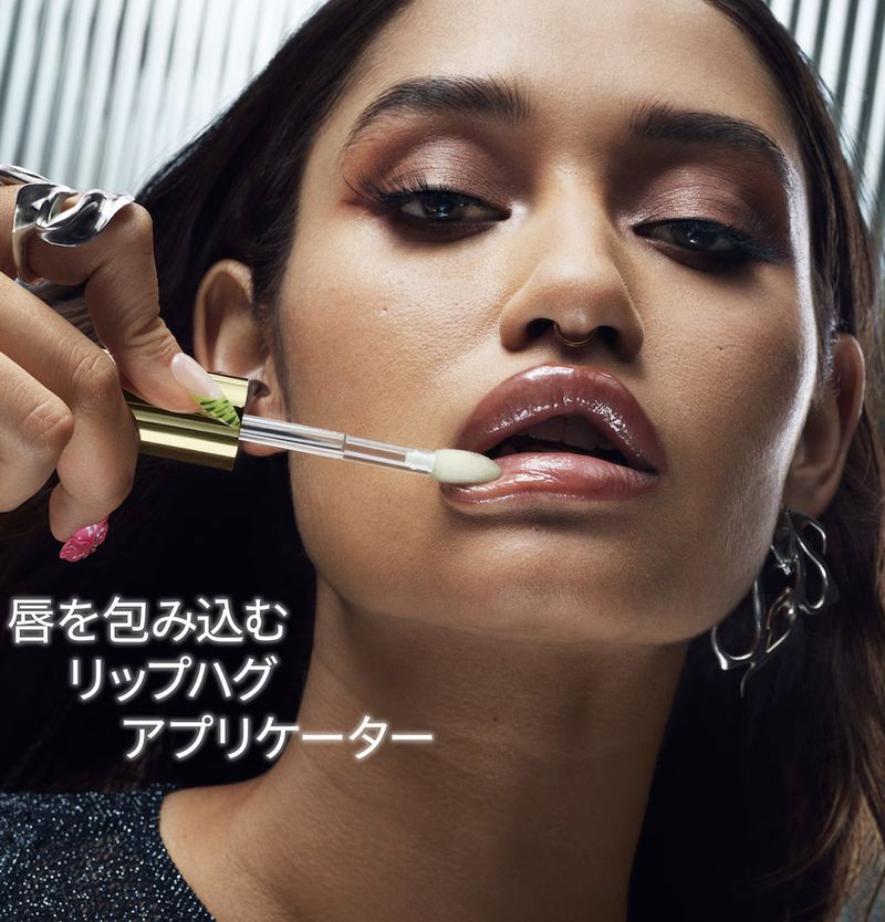Close-up shot of a woman with glossy lips, an applicator, and jewelry, against a silver background, showcasing a beauty product.