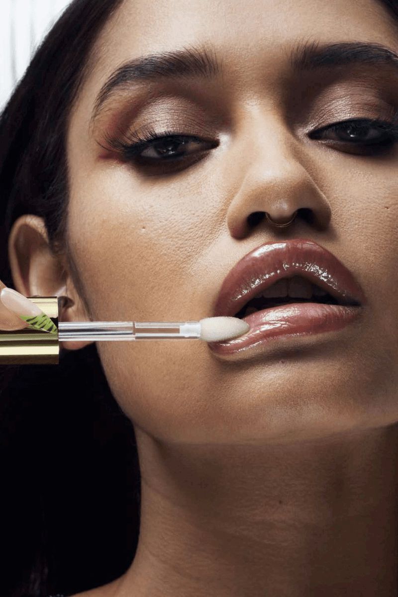 Close-up shot of a young adult female applying clear lip gloss with an applicator, showcasing glossy lips, makeup and nail art details.