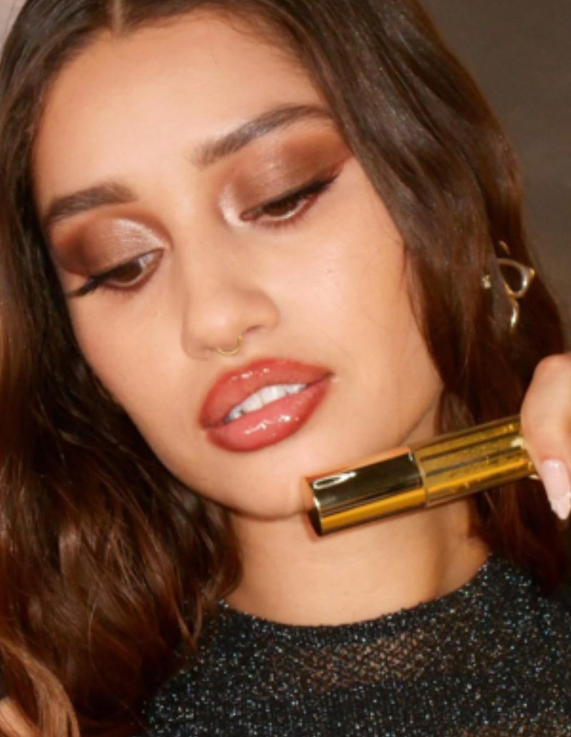 Close-up of a woman with brown hair holding a gold-tone cosmetic tube, glossy lips, and full makeup. Beauty and cosmetic product shot.