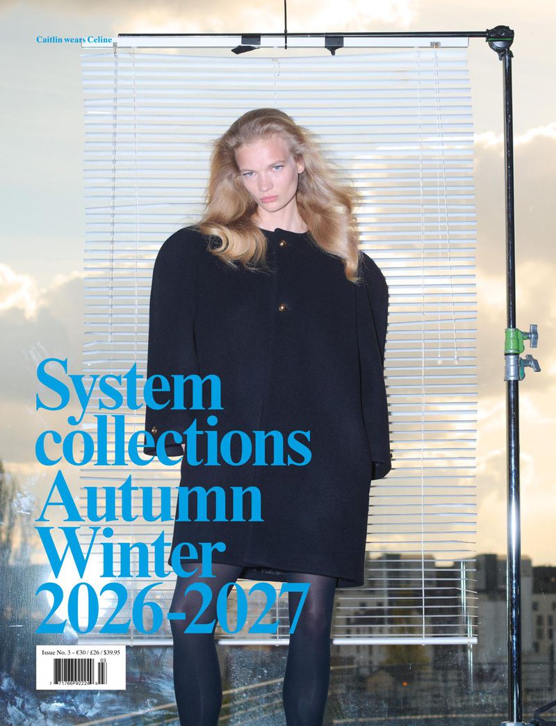 Model with long blonde hair wearing a black coat and tights standing in front of a white window blind backdrop