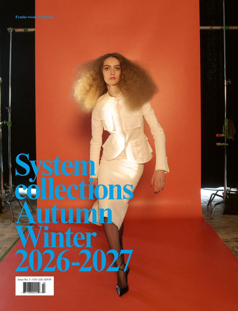 Model in white jacket and skirt standing in front of an orange seamless background with studio lighting.