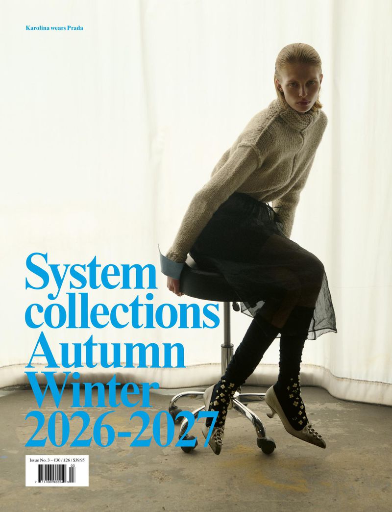 Model seated on a rolling stool wearing a textured turtleneck sweater and sheer skirt against white curtains.