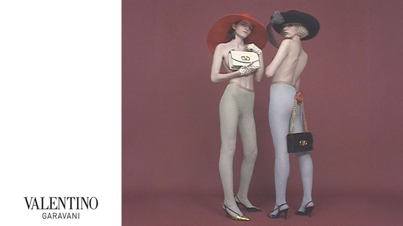 Two fashion models pose in wide-brimmed hats, pantyhose, and kitten heels while holding purses against a mauve background in a studio shot.