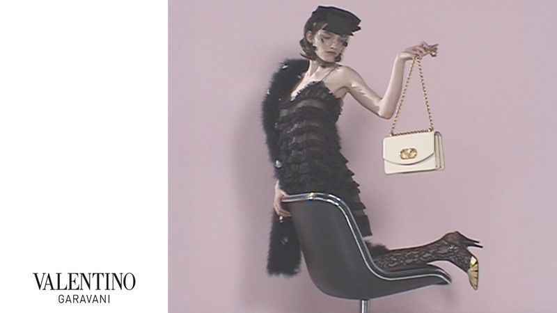 Fashion editorial shot of a woman in black dress and hat sitting on a chair, holding a handbag with the Valentino Garavani logo.