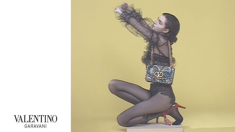 A woman poses in a black sheer outfit, kneeling in front of a yellow backdrop, modeling a patterned handbag and heeled shoes for a fashion shoot.