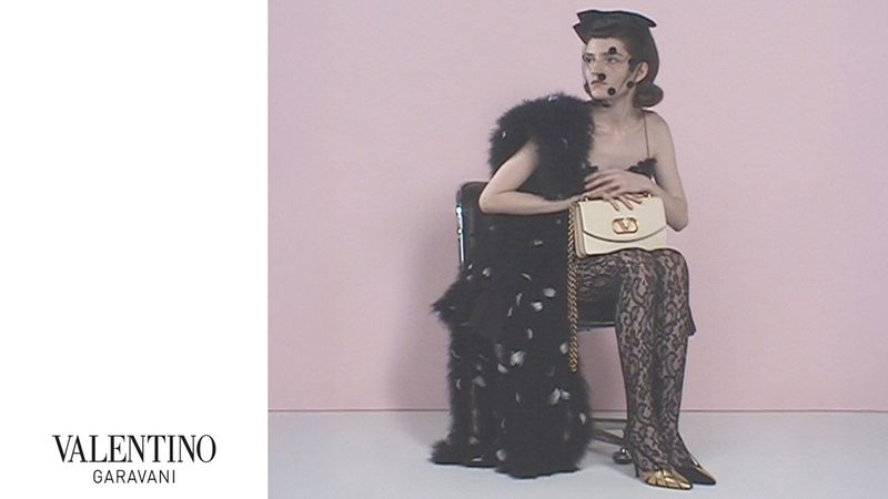 Woman sits in black dress with feathers, lace tights, metallic heels, and a Valentino handbag against a light pink background.
