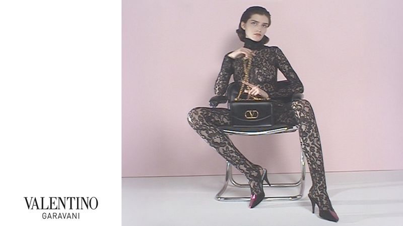 A woman in a black lace catsuit poses while seated on a chair, holding a black purse, against a light pink studio backdrop.