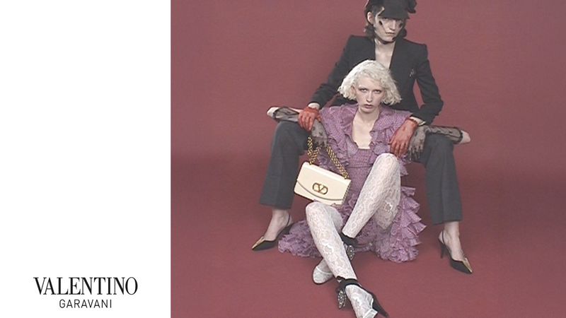 Two fashion models pose indoors against a maroon backdrop. The model in front wears a purple dress with lace stockings and a white purse.