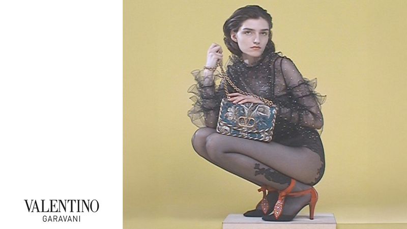 Woman poses crouching with a Valentino Garavani handbag. She wears a sheer black top, patterned hosiery, and high heels against a yellow backdrop.