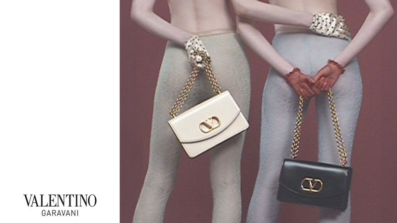 Valentino Garavani brand campaign features two models holding Valentino handbags against a mauve backdrop in a studio setting.
