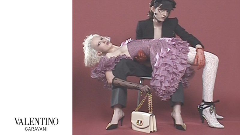 Two people in a fashion editorial: one woman in a ruffled dress being held by another person seated on a stool with a purse.