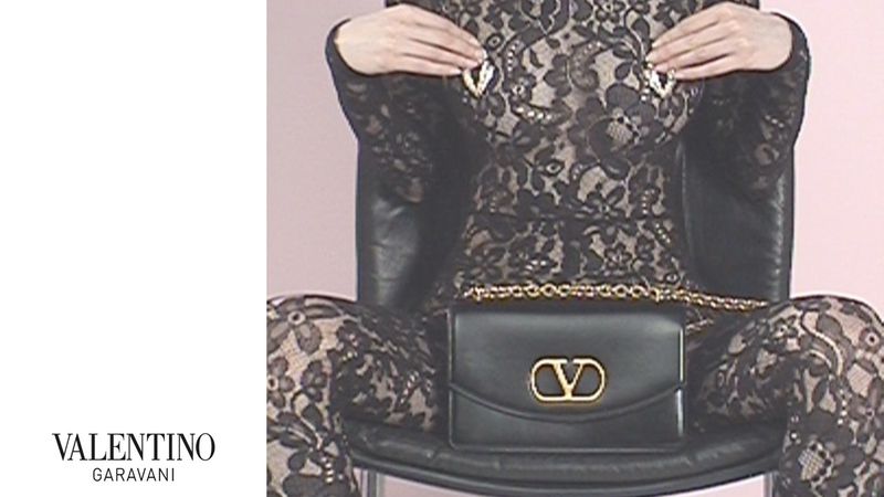A person is seated in a chair wearing a lace jumpsuit, holding jewelry near their shoulders, and a Valentino Garavani purse.