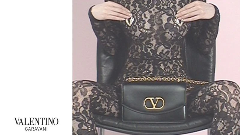 A person is seated in a chair wearing a lace jumpsuit, holding jewelry near their shoulders, and a Valentino Garavani purse.