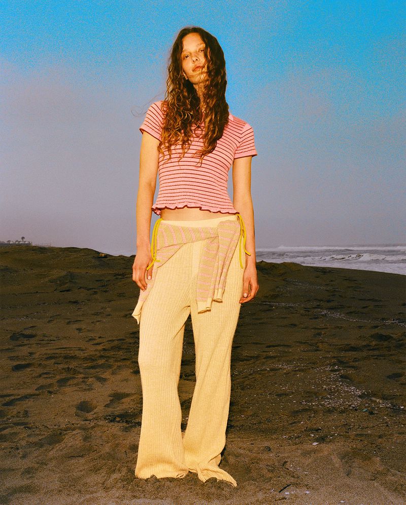 Model wearing a pink striped top and pale yellow trousers standing on a sandy beach against a blue sky.