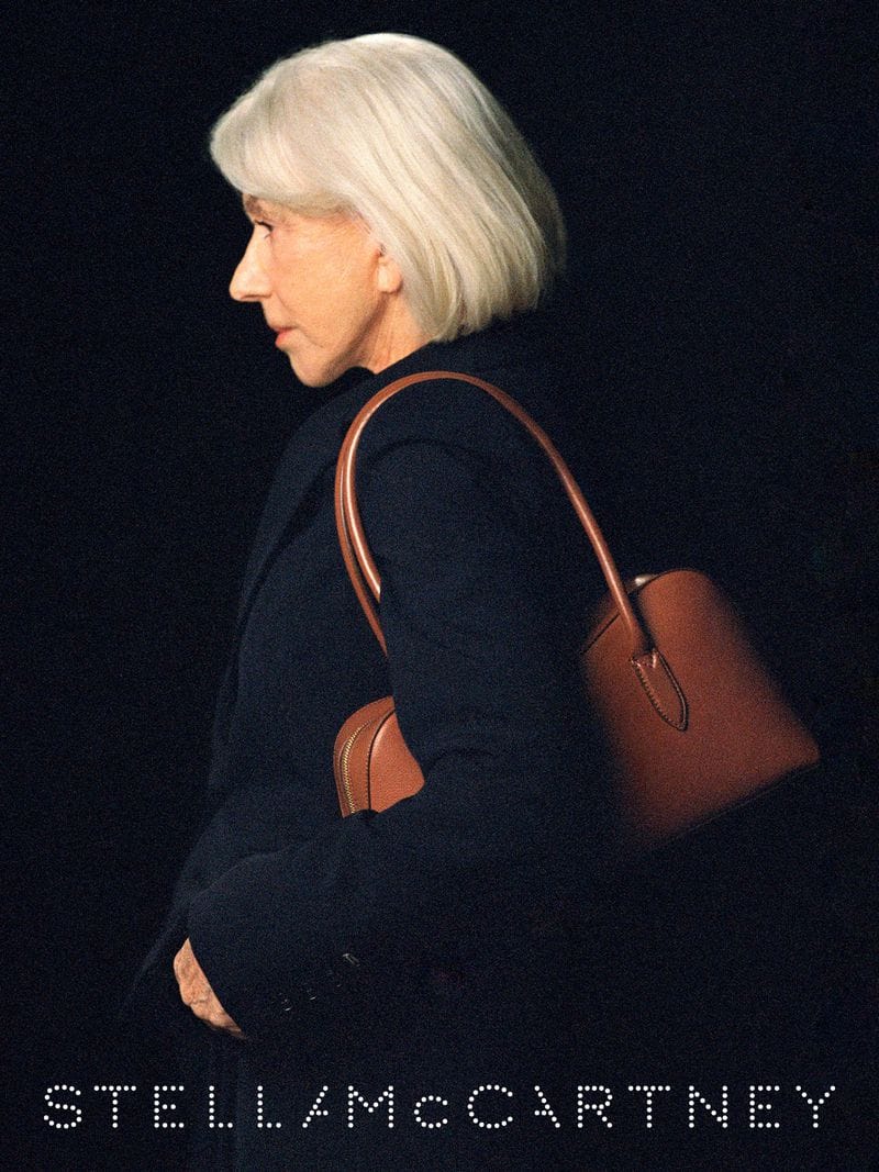 Helen Mirren wearing dark coat holding a brown leather shoulder bag against black background for Stella McCartney