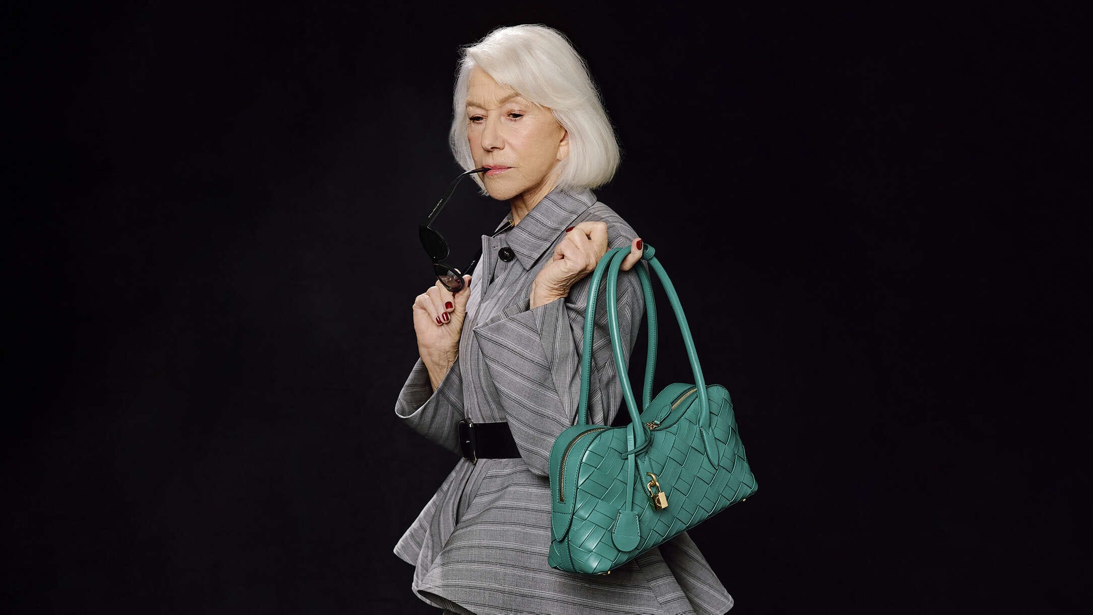 Helen Mirren wearing grey belted jacket holding a teal leather handbag against a dark background