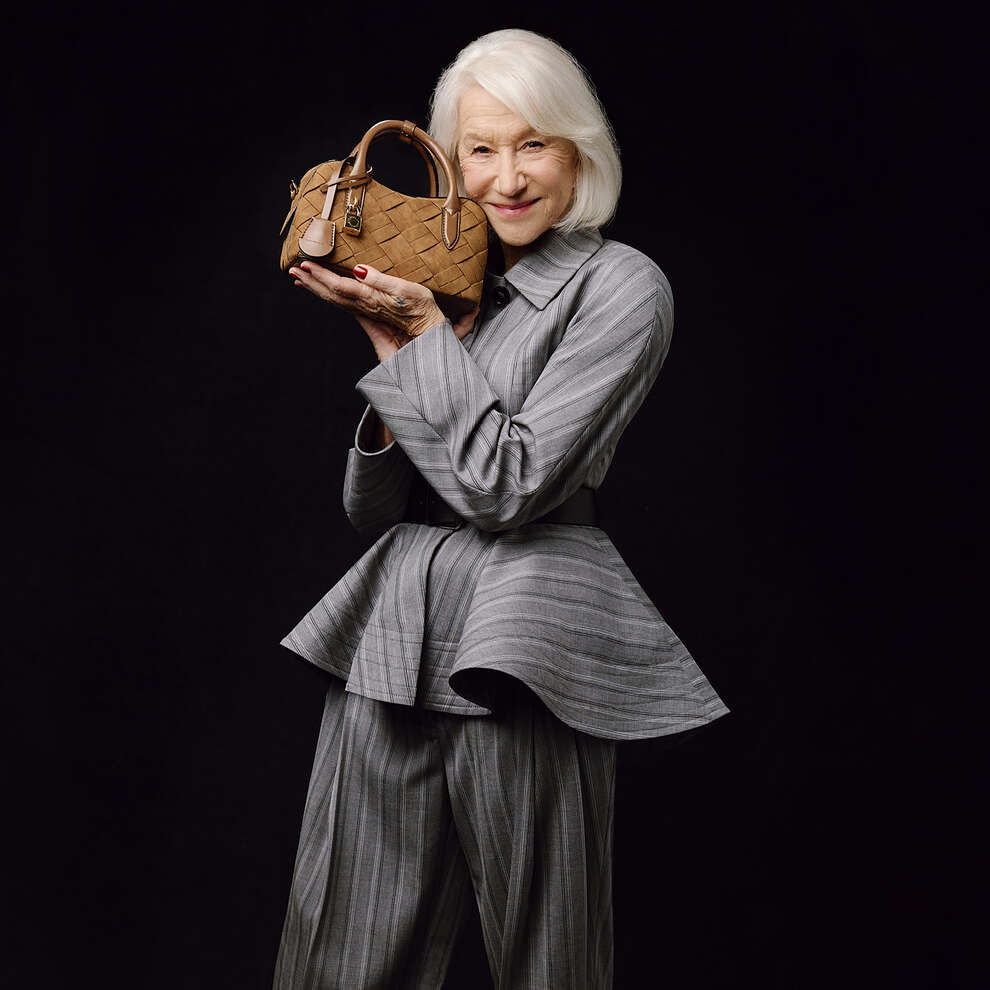 Helen Mirren wearing a grey striped suit holding a brown woven handbag against a black seamless background