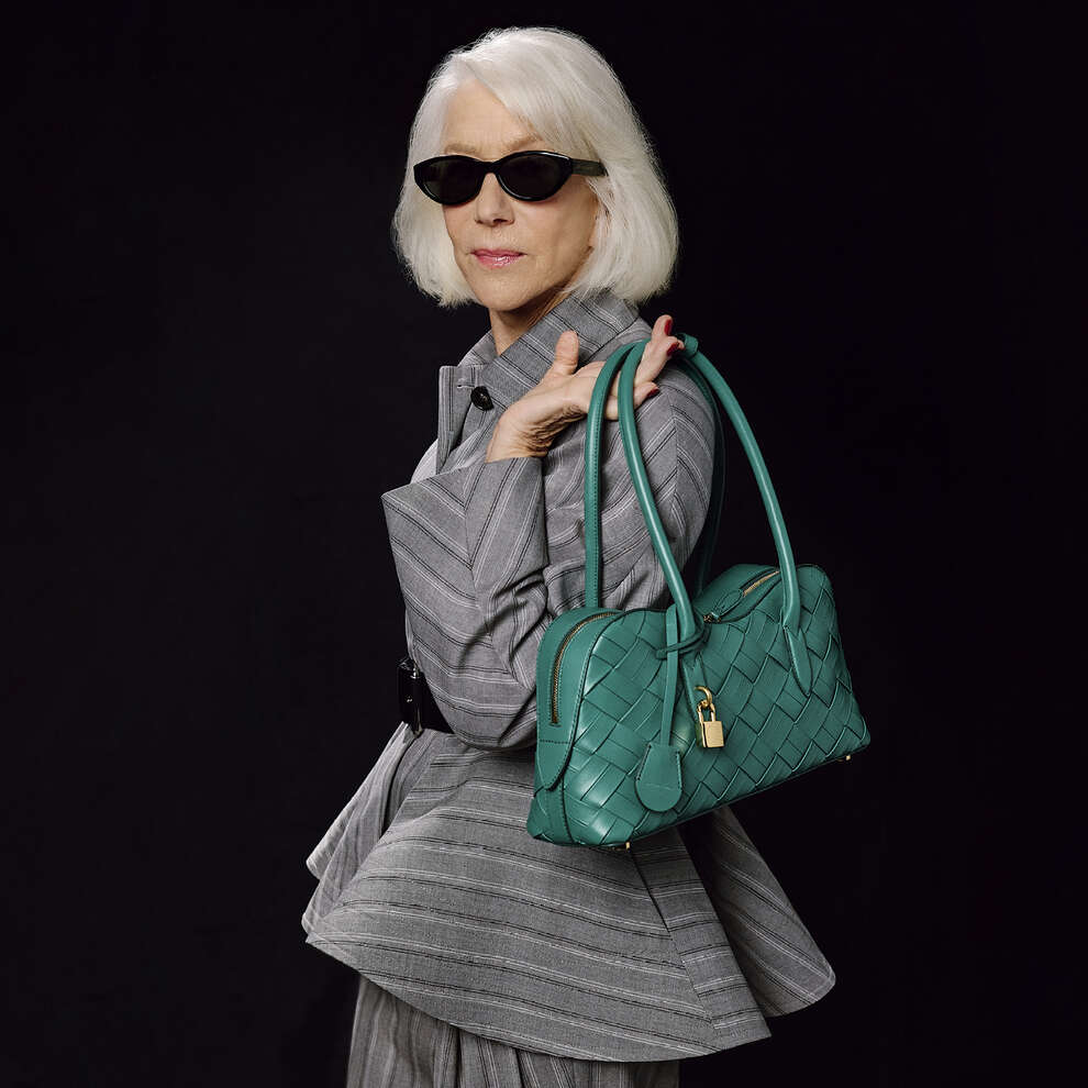 Helen Mirren wearing dark sunglasses and a grey patterned suit holding a green woven handbag against a black background
