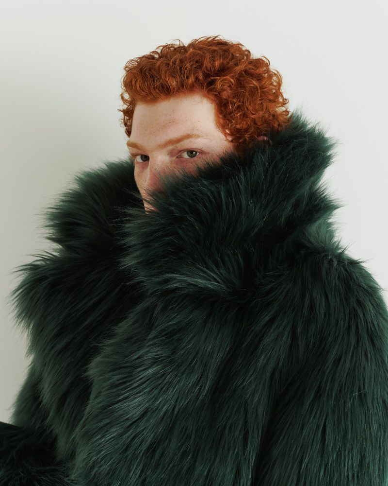 Close-up studio portrait of a red-haired male adult wearing a voluminous, faux fur coat in a dark green hue against a plain white background.
