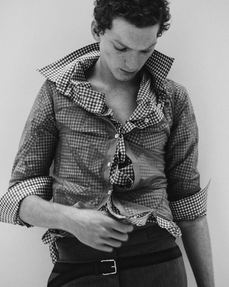 Monochrome portrait of a young adult male with curly hair, wearing an unbuttoned checkered shirt and dark pants with a belt in an indoor studio.