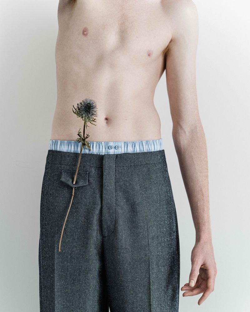 Torso shot of shirtless adult male wearing grey trousers with a thistle on his stomach in a photo studio against a white backdrop.