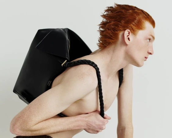 Profile shot of an adult male model with red hair wearing a large black leather backpack against a plain white backdrop in a photo studio.
