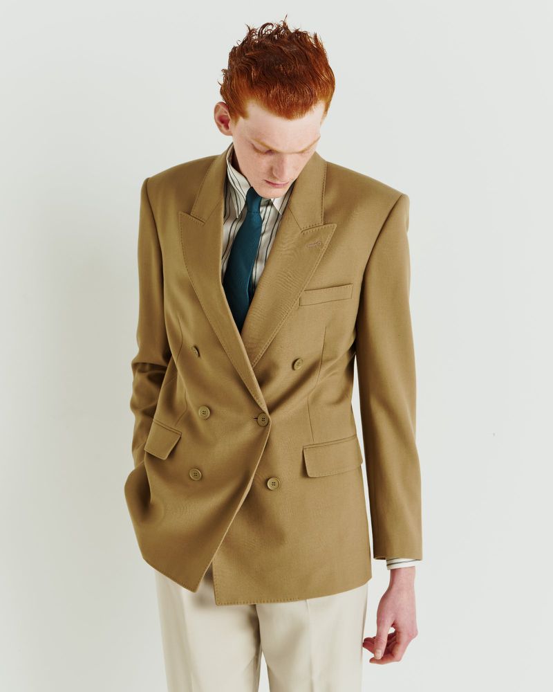 Red-haired male model in a double-breasted tan blazer with a green tie stands in a light studio wearing cream trousers.