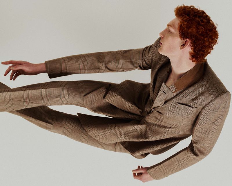 Red-haired man in a plaid suit lying horizontally against a white background, fashion pose, formal wear, studio lighting. 