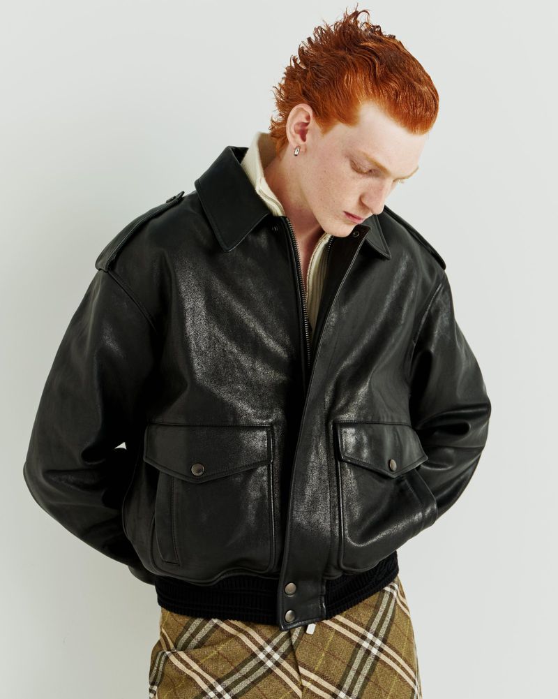 Adult male with red hair poses in a black leather jacket with pockets and buttons against a white background in a studio shot.