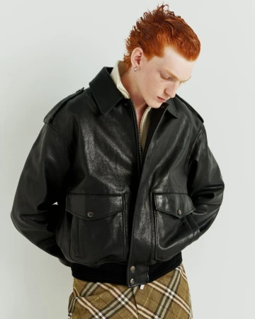 Adult male with red hair poses in a black leather jacket with pockets and buttons against a white background in a studio shot.