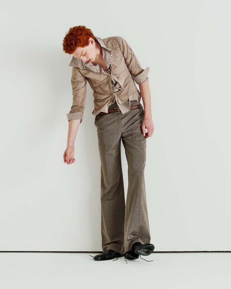 Model with red hair wearing a patterned shirt and brown trousers against a white seamless studio backdrop